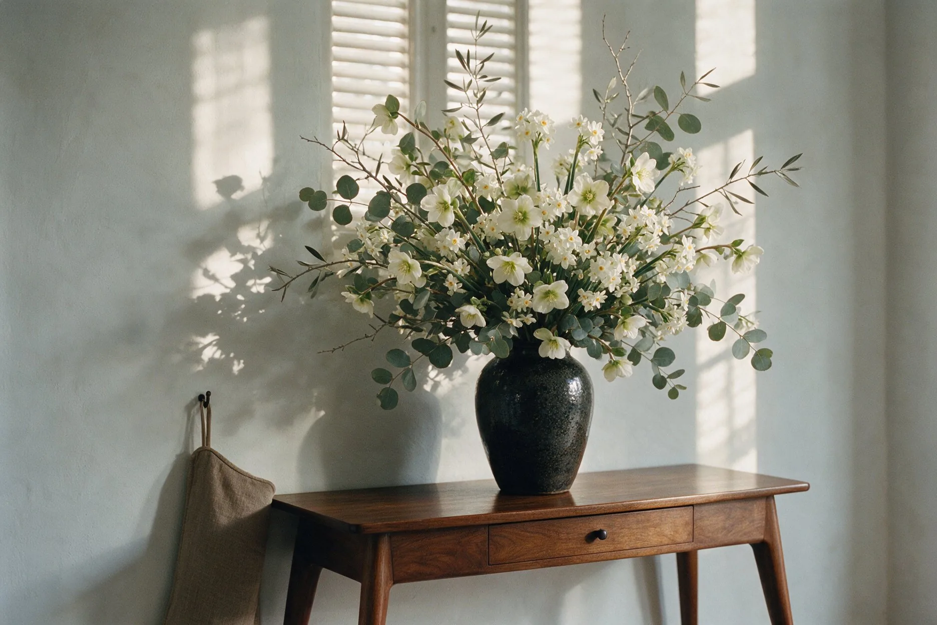 White hellebores, narcissus, eucalyptus and bare olive in a dark glazed vase on a walnut console, morning light on a lime-washed wall